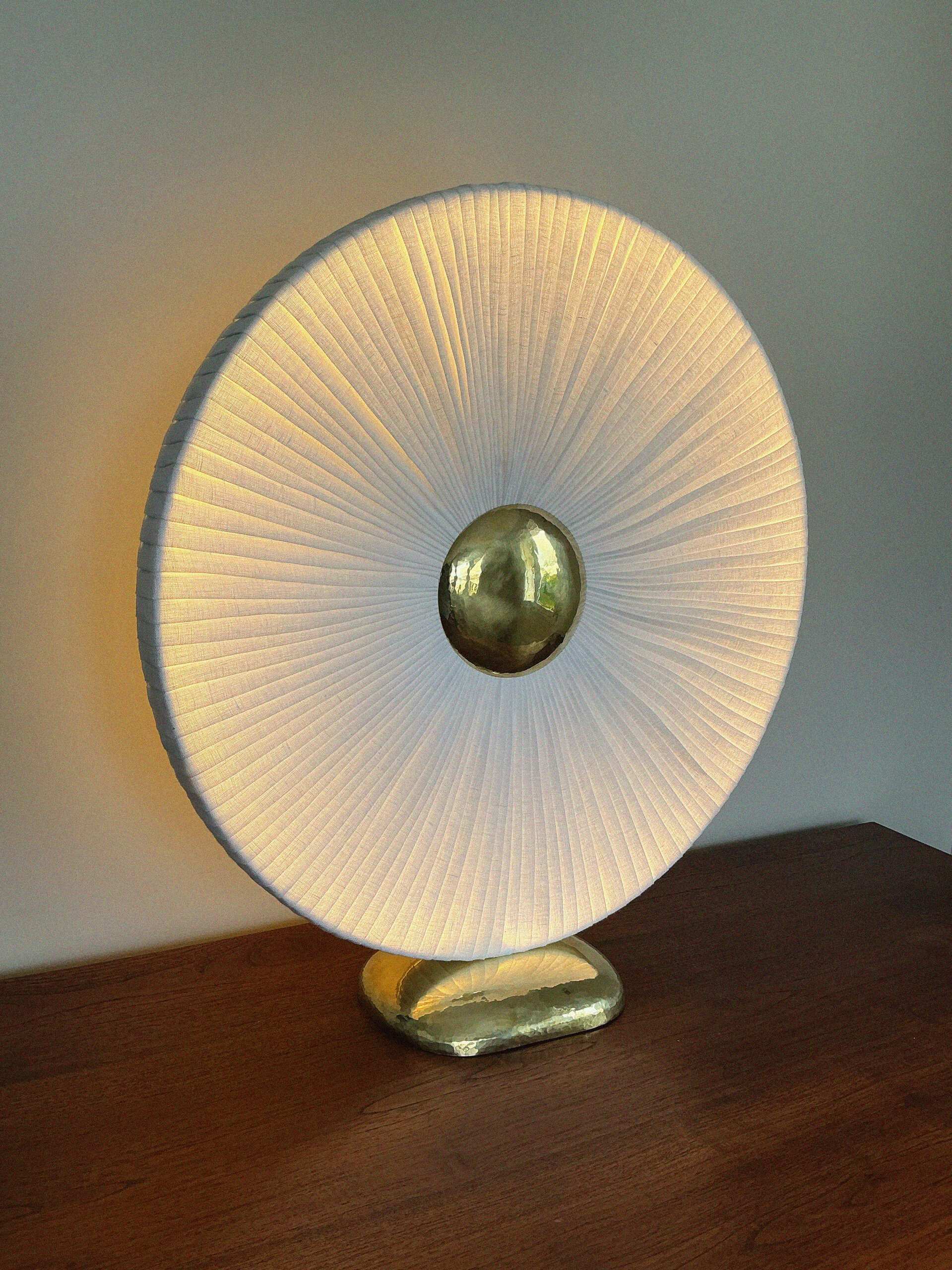 Iris allumée : disque de lin plissé diffusant une lumière chaude, cœur et socle en laiton martelé, posée sur un meuble en bois.